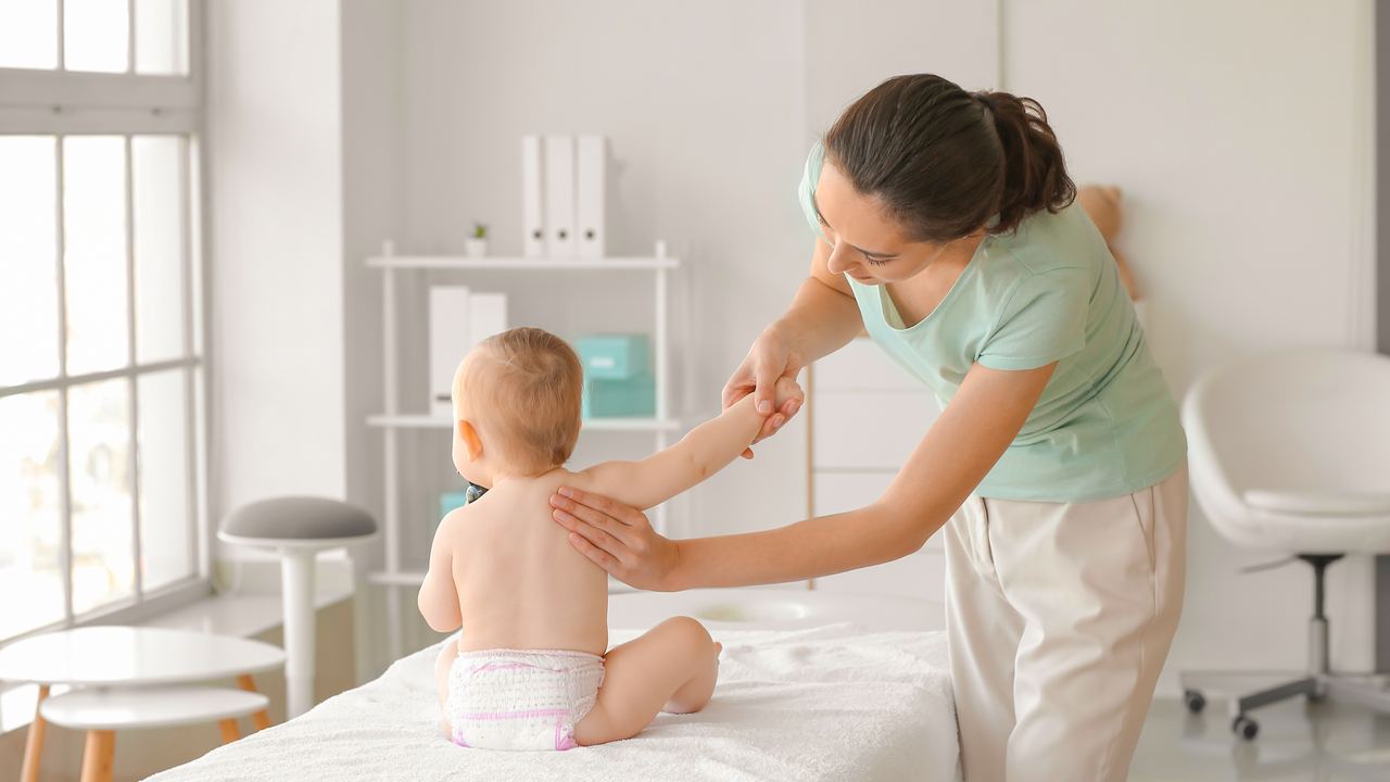 Therapeutin stützt ein sitzendes Baby auf der Liege und führt seinen Arm