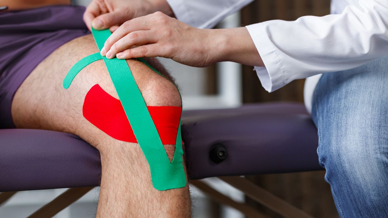 Rotes und grünes Kinesio-Tape auf einem Knie, Hände kleben den letzten Streifen