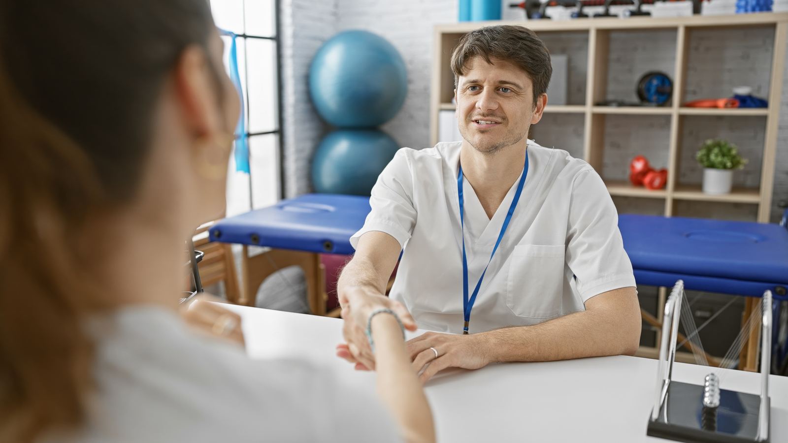 Therapeut im Gespräch mit einer Patientin vor der Behandlung, dahinter Behandlungsliege und Übungsgeräte